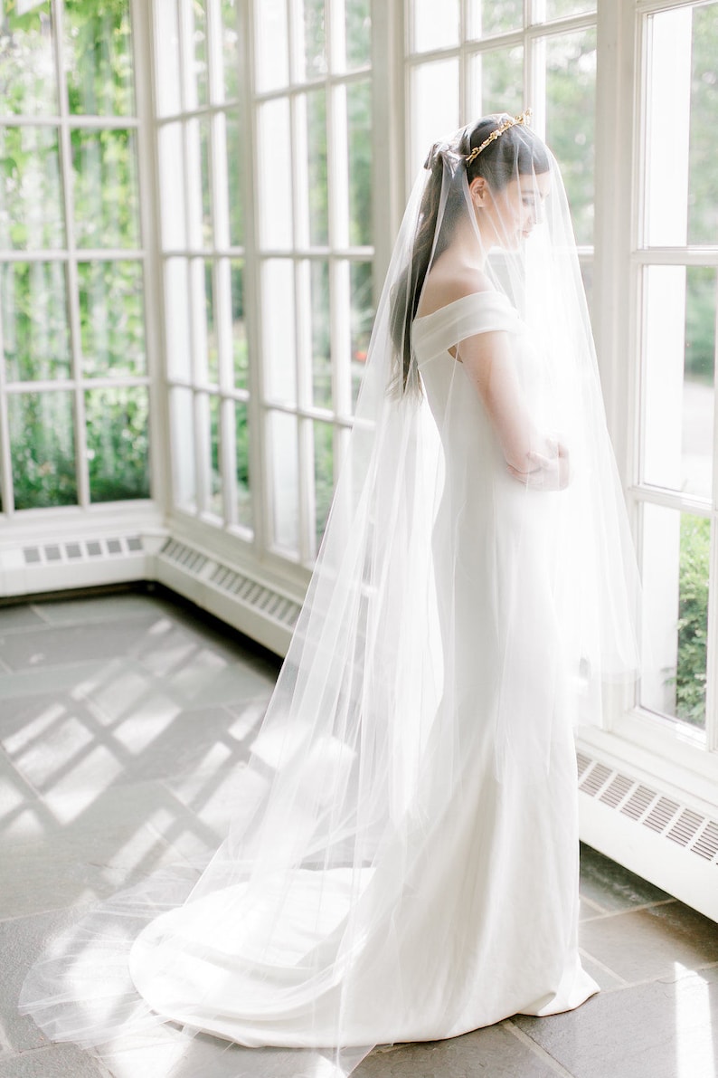 Puede incluir: Una novia con un vestido de novia blanco con un velo largo y una corona dorada. Est&aacute; de pie en una habitaci&oacute;n soleada con grandes ventanas.