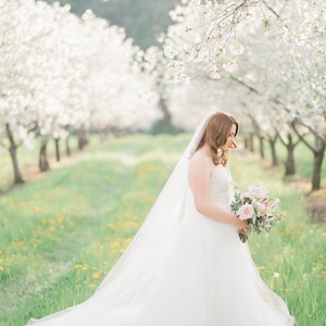 Puede incluir: Una novia con un vestido de novia blanco y velo sostiene un ramo de flores rosas y blancas en un campo de hierba verde y flores blancas.