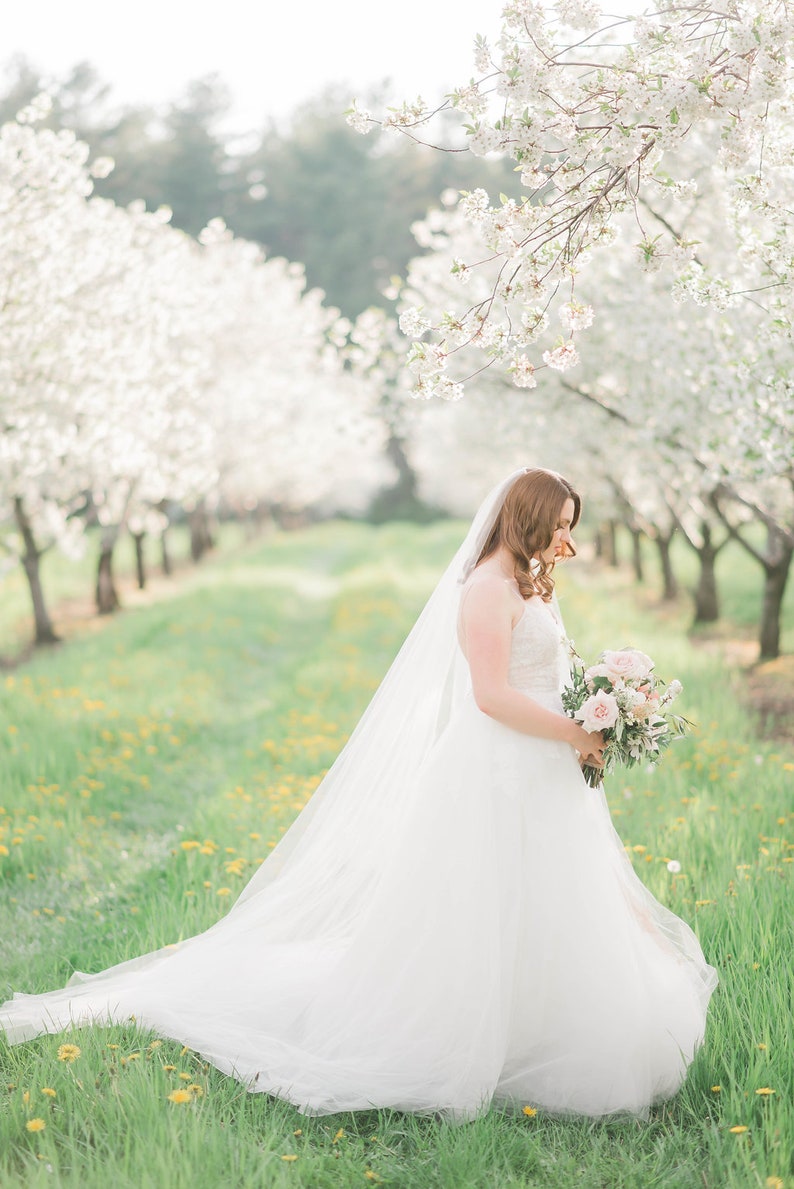 Puede incluir: Una novia con un vestido de novia blanco y velo sostiene un ramo de flores rosas y blancas en un campo de hierba verde y flores blancas.