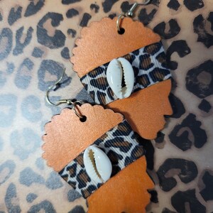 May include: A pair of orange wooden earrings in the shape of a woman's profile. Each earring features a band of leopard print fabric and a white seashell. The earrings have gold-colored ear wires and are displayed on a leopard print background.