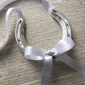 May include: A silver horseshoe with a white satin ribbon tied around it. The horseshoe has the text "2 KERCKHAER" engraved on it.
