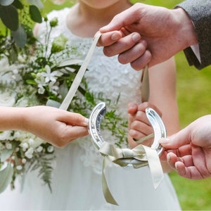 May include: A silver horseshoe with a white satin ribbon tied in a bow. The horseshoe is being held by two people, one of whom is wearing a white dress.
