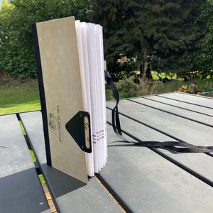 May include: A beige, closed, tri-guard file with a black strap and a black metal clasp. The file is labeled "Tri-Guard File" and has a black label with the text "Go 18" on it.