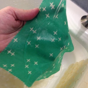 Peut inclure: Un emballage alimentaire r&eacute;utilisable vert avec un motif de croix blanches est rinc&eacute; sous l'eau courante dans un &eacute;vier blanc.