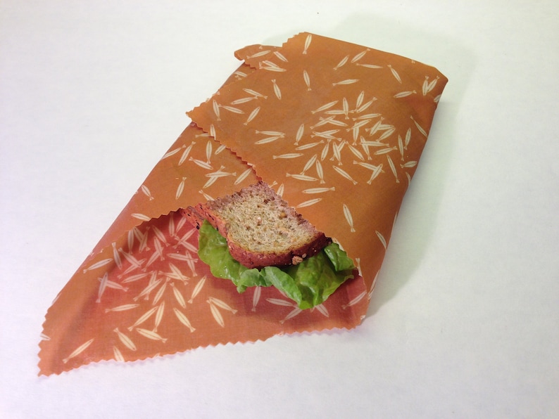 Peut inclure: Un sandwich envelopp&eacute; dans une pellicule alimentaire r&eacute;utilisable brune avec un motif de poisson blanc. Le sandwich est compos&eacute; de pain complet, de laitue et d'une tranche de fromage.