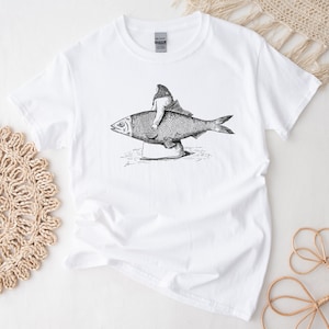 Könnte beinhalten: Weißes T-Shirt mit einer Schwarzweißillustration einer Person, die auf einem Fisch reitet.