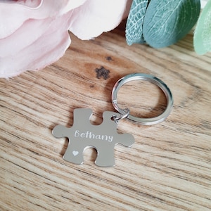 May include: Silver puzzle piece keyring with the name Bethany engraved on it. The keyring has a small heart engraved on it.