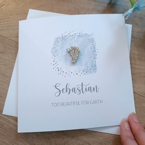 Peut inclure: Une carte bleu clair avec un charme de pied de b&eacute;b&eacute; argent&eacute; avec des strass. La carte dit "S&eacute;bastien Trop beau pour la Terre".
