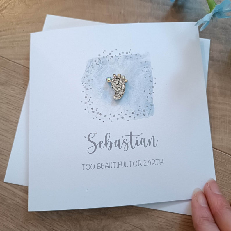 Peut inclure: Une carte bleu clair avec un charme de pied de b&eacute;b&eacute; argent&eacute; avec des strass. La carte dit "S&eacute;bastien Trop beau pour la Terre".
