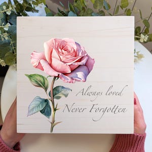 May include: A square wooden sign featuring a pink rose illustration with green leaves. The text "Always loved, Never Forgotten" is written in script. The background is white with a wood grain effect.