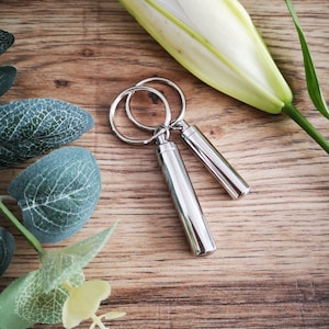 May include: Two silver keychains with small cylindrical containers attached. The keychains are on a wooden surface with green leaves and a white flower.