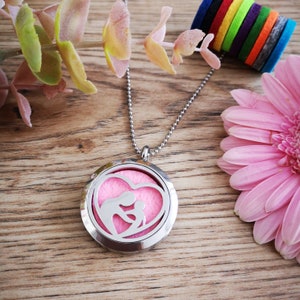 Könnte beinhalten: Eine silberne Halskette mit einem herzförmigen Anhänger, der ein ausgeschnittenes Design einer Mutter und eines Kindes zeigt. Der Anhänger ist offen und zeigt ein rosa Filzpolster im Inneren.