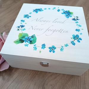 May include: A light-colored wooden box with a hinged lid. The lid is decorated with a wreath of blue flowers and green leaves, with the words "Always loved, Never forgotten" in script.