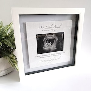 Pode incluir: Impressão de ultrassom emoldurada em branco com o texto "Our Little Angel 8th January 2025" e a citação "Too Beautiful for Earth". A imagem mostra uma ultrassonografia em tons de cinza de um bebê.