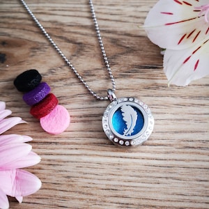 May include: A silver chain necklace with a round pendant featuring a blue feather design and clear rhinestones. The pendant is attached to a silver chain with a lobster clasp. The necklace is displayed on a wooden surface with colorful felt circles.