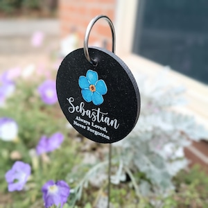 Könnte beinhalten: Ein schwarzer, runder Gartenstecker mit einer silbernen Metallschlaufe. Er zeigt ein blaues Blumendesign und den Namen "Sebastian" mit dem Text "Always Loved, Never Forgotten" in weißer Schrift. Der Stecker befindet sich in einem Garten.