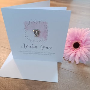 Peut inclure: Une carte blanche avec un fond aquarelle rose et une empreinte de pied en paillettes argent&eacute;es avec un strass. La carte dit "Amelia Grace" et "M&ecirc;me les plus petits pieds ont le pouvoir de laisser des empreintes &eacute;ternelles sur ce monde."