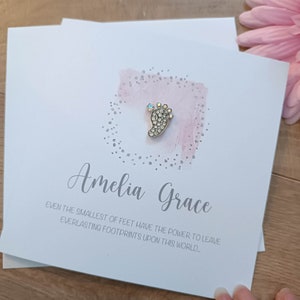 Peut inclure: Une carte blanche avec un fond aquarelle rose et un charme de pied de b&eacute;b&eacute; argent&eacute; avec des strass. La carte dit "Amelia Grace" et "M&ecirc;me les plus petits pieds ont le pouvoir de laisser des empreintes &eacute;ternelles sur ce monde."