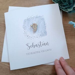 Peut inclure: Une carte blanche avec un charme de pied de b&eacute;b&eacute; argent&eacute; avec des strass. La carte porte le nom "Sebastian" &eacute;crit en cursive et le texte "Trop beau pour la terre".