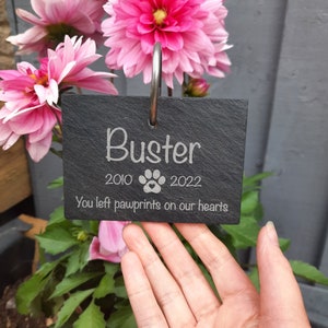 May include: Black slate memorial plaque engraved with the name "Buster", dates "2010" and "2022", a paw print with a heart, and the text "You left pawprints on our hearts".