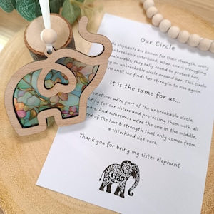 May include: Wooden elephant ornament with a floral stained-glass design. The elephant has a light wood frame and a colorful interior with pink, blue, and green hues. A white ribbon is attached for hanging. A card with text and an elephant illustration is next to the ornament.