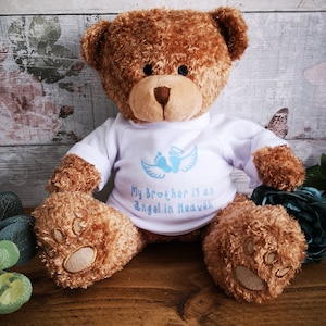 Könnte beinhalten: Ein brauner Teddybär, der ein weißes Shirt mit blauen Engelsflügeln und Fußabdrücken trägt. Auf dem Shirt steht "Mein Bruder ist ein Engel im Himmel."