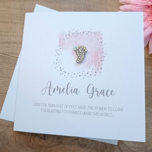 Peut inclure: Une carte blanche avec un fond aquarelle rose et des paillettes argent&eacute;es. Un charme de pied de b&eacute;b&eacute; argent&eacute; avec des strass est attach&eacute; &agrave; la carte. La carte indique "Amelia Grace" et "M&ecirc;me les plus petits pieds ont le pouvoir de laisser des empreintes &eacute;ternelles sur ce monde."