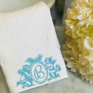 a hand towel embroidered with angels and a monogram letter B. White flowers by the side of the towel