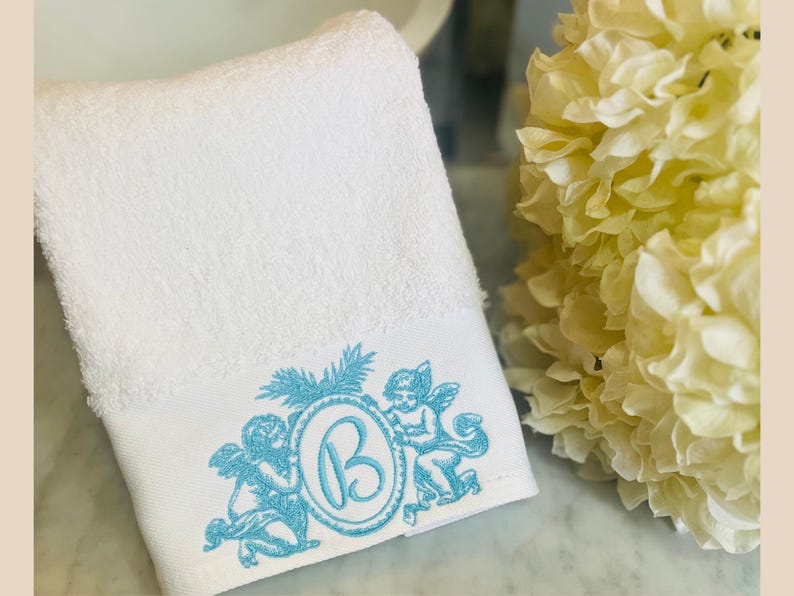a hand towel embroidered with angels and a monogram letter B. White flowers by the side of the towel