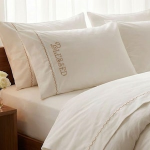 May include: Cream-coloured bedding set with embroidered details. The pillowcases feature the word "Blessed" in a script font. The set includes pillowcases and a duvet cover, all with a decorative stitched edge. A vase of white roses sits on a wooden bedside table.