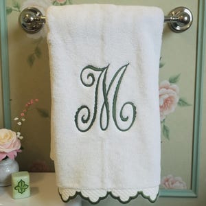 Puede incluir: Toalla de baño blanca con una letra M bordada en verde. La toalla tiene un borde festoneado.