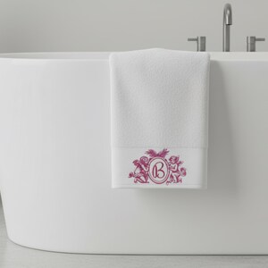 A white bath towel hanging on the side of a bath tub. The towel is embroidered with two cherub angels and monogrammed with the letter B. The embroidery is pink.