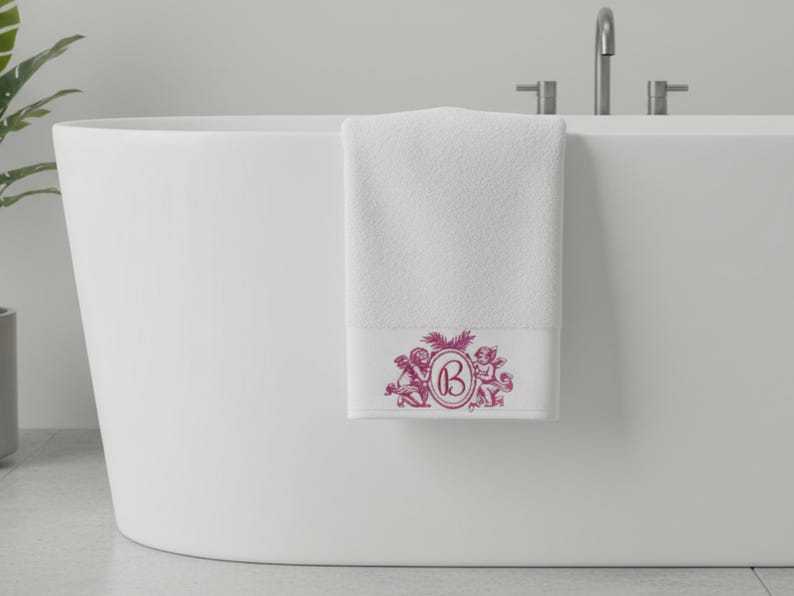 A white bath towel hanging on the side of a bath tub. The towel is embroidered with two cherub angels and monogrammed with the letter B. The embroidery is pink.