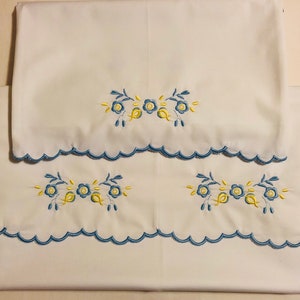 Könnte beinhalten: Zwei weiße Kissenbezüge mit blauen gewellten Rändern und gestickten gelben und blauen Blumenmustern.