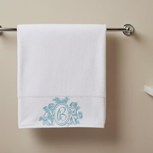 A bath  towel hanging on a rack. The bath towel is embroidered at the bottom with two cherub angels and monogrammed with the letter B.  There is a plant by the side of the towel.