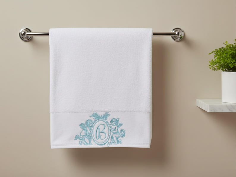 A bath  towel hanging on a rack. The bath towel is embroidered at the bottom with two cherub angels and monogrammed with the letter B.  There is a plant by the side of the towel.