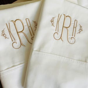 May include: Two ivory-colored bed sheets with gold embroidered monograms. The monogram features the letter "R" in an elegant script, with decorative flourishes and the number "1804" embroidered on each side. The sheets are folded.