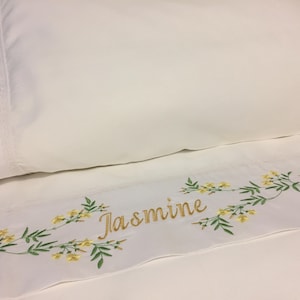 May include: White bed linen set featuring embroidered floral and leaf patterns in yellow and green. The name "Jasmine" is embroidered in gold script. A floral initial is embroidered on the pillowcase.