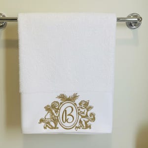 Puede incluir: Toalla de ba&ntilde;o blanca con un dise&ntilde;o bordado en oro que presenta dos querubines que flanquean un &oacute;valo con la letra "B". La toalla est&aacute; colgada de una barra cromada. El dise&ntilde;o est&aacute; en la parte inferior de la toalla.