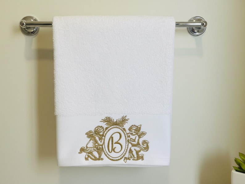 Puede incluir: Toalla de ba&ntilde;o blanca con un dise&ntilde;o bordado en oro que presenta dos querubines que flanquean un &oacute;valo con la letra "B". La toalla est&aacute; colgada de una barra cromada. El dise&ntilde;o est&aacute; en la parte inferior de la toalla.