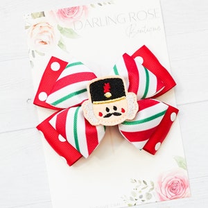 May include: A festive hair bow featuring a red, white, and green striped pattern with a red polka dot accent. The bow is adorned with an embroidered nutcracker face. The background features the text "Darling Rose Boutique".