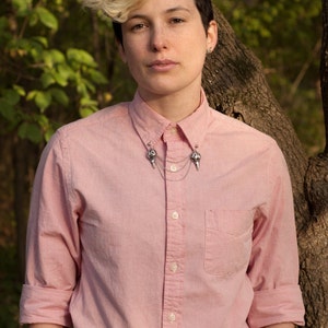 May include: A person wearing a pink button-down shirt with a silver collar pin. The collar pin has two skull-shaped charms connected by a chain.
