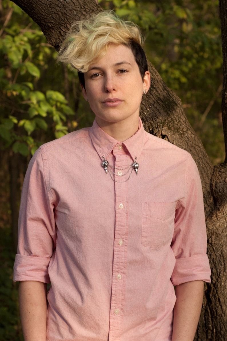 May include: A person wearing a pink button-down shirt with a silver collar pin. The collar pin has two skull-shaped charms connected by a chain.