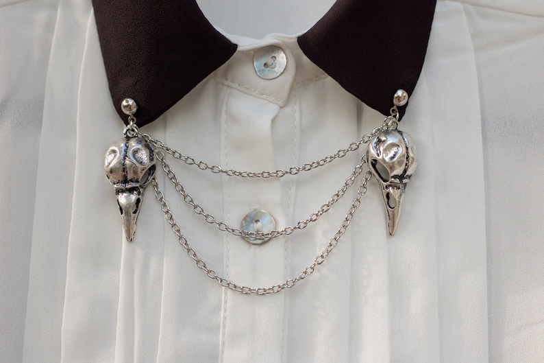 May include: A silver skull pendant with a chain necklace. The pendant is attached to a black collar on a white shirt.