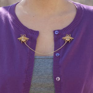 May include: A gold-colored bee cardigan clip with a chain connecting two bee-shaped brooches. The clip is attached to a purple cardigan over a gray top. The cardigan has matching purple buttons.