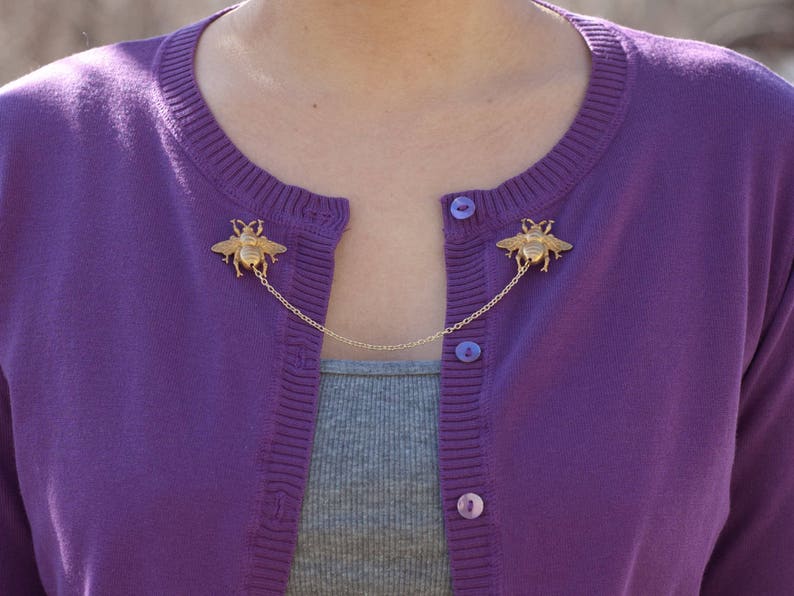 May include: A gold-colored bee cardigan clip with a chain connecting two bee-shaped brooches. The clip is attached to a purple cardigan over a gray top. The cardigan has matching purple buttons.