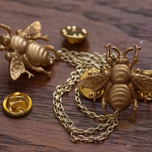 May include: Gold-colored bee-shaped lapel pins with intricate details, including wings and antennae. One pin is connected to another with a delicate chain. Two additional pin backs are also visible. The pins are displayed on a dark wooden surface.
