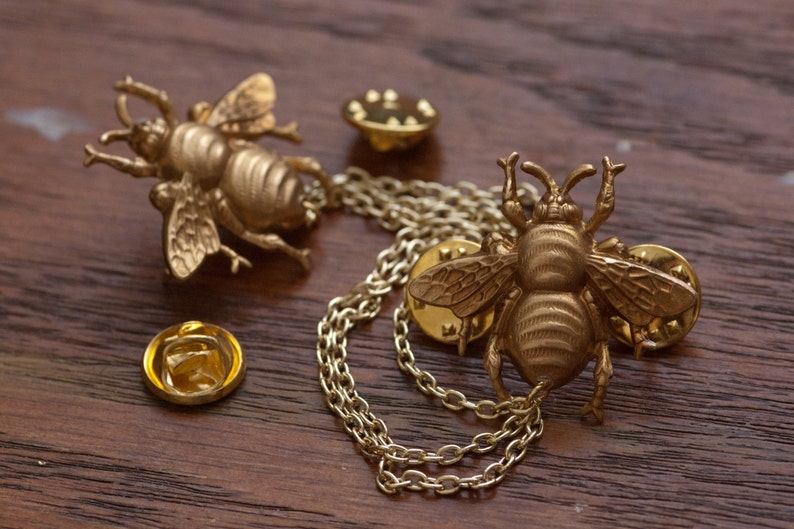 May include: Gold-colored bee-shaped lapel pins with intricate details, including wings and antennae. One pin is connected to another with a delicate chain. Two additional pin backs are also visible. The pins are displayed on a dark wooden surface.