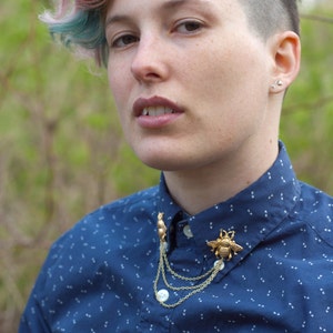 May include: A blue button-down shirt with a white pattern. The shirt features a gold-colored bee and squirrel brooch with dangling chains. The person has short, multi-colored hair and small earrings.