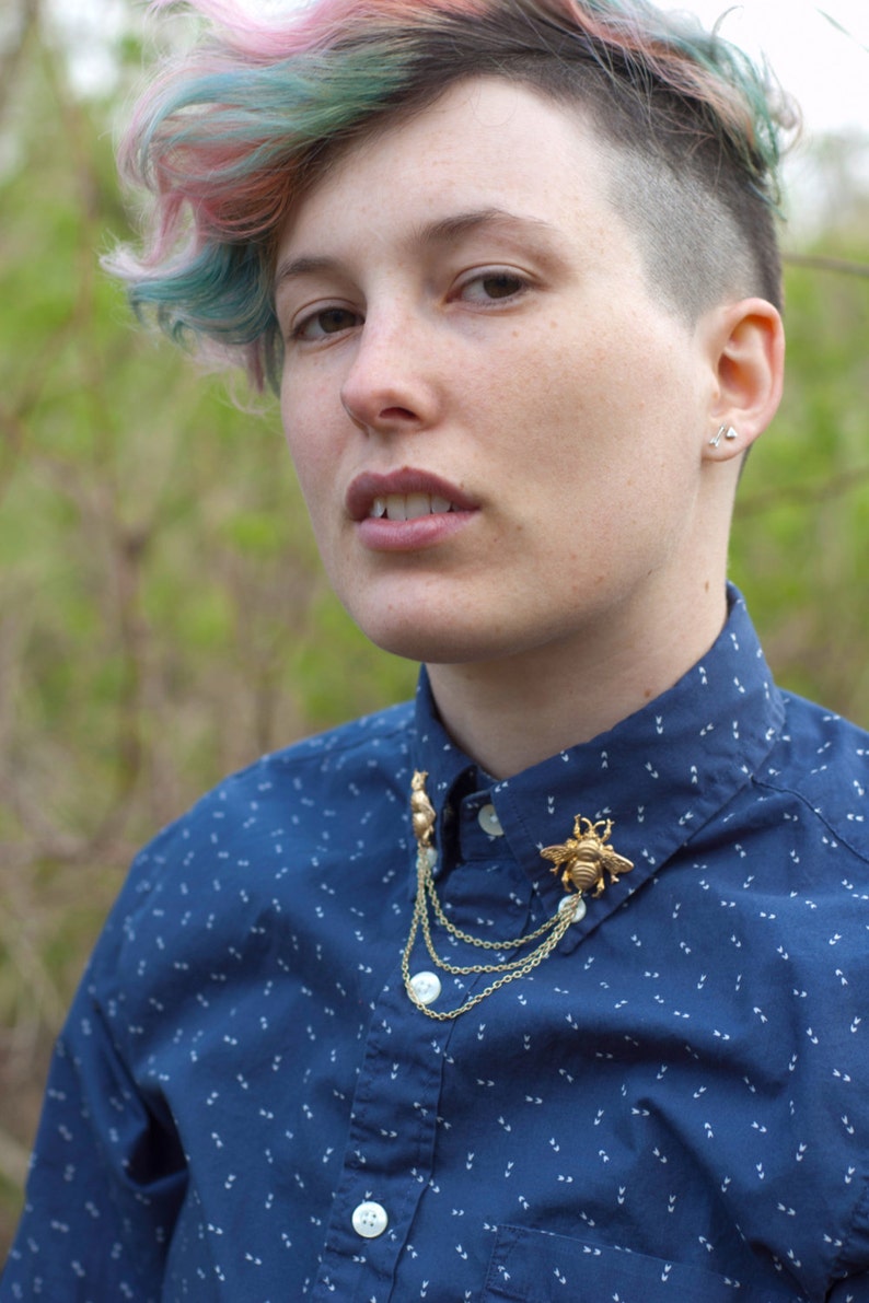 May include: A blue button-down shirt with a white pattern. The shirt features a gold-colored bee and squirrel brooch with dangling chains. The person has short, multi-colored hair and small earrings.